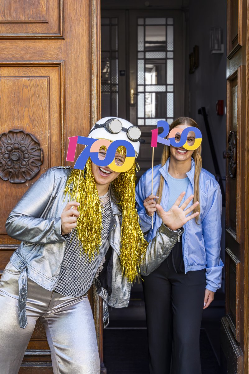 Zwei Frauen feiern mit Party-Accessoires und 1700-Schildern vor einer Holztür bei einer festlichen Firmenveranstaltung.